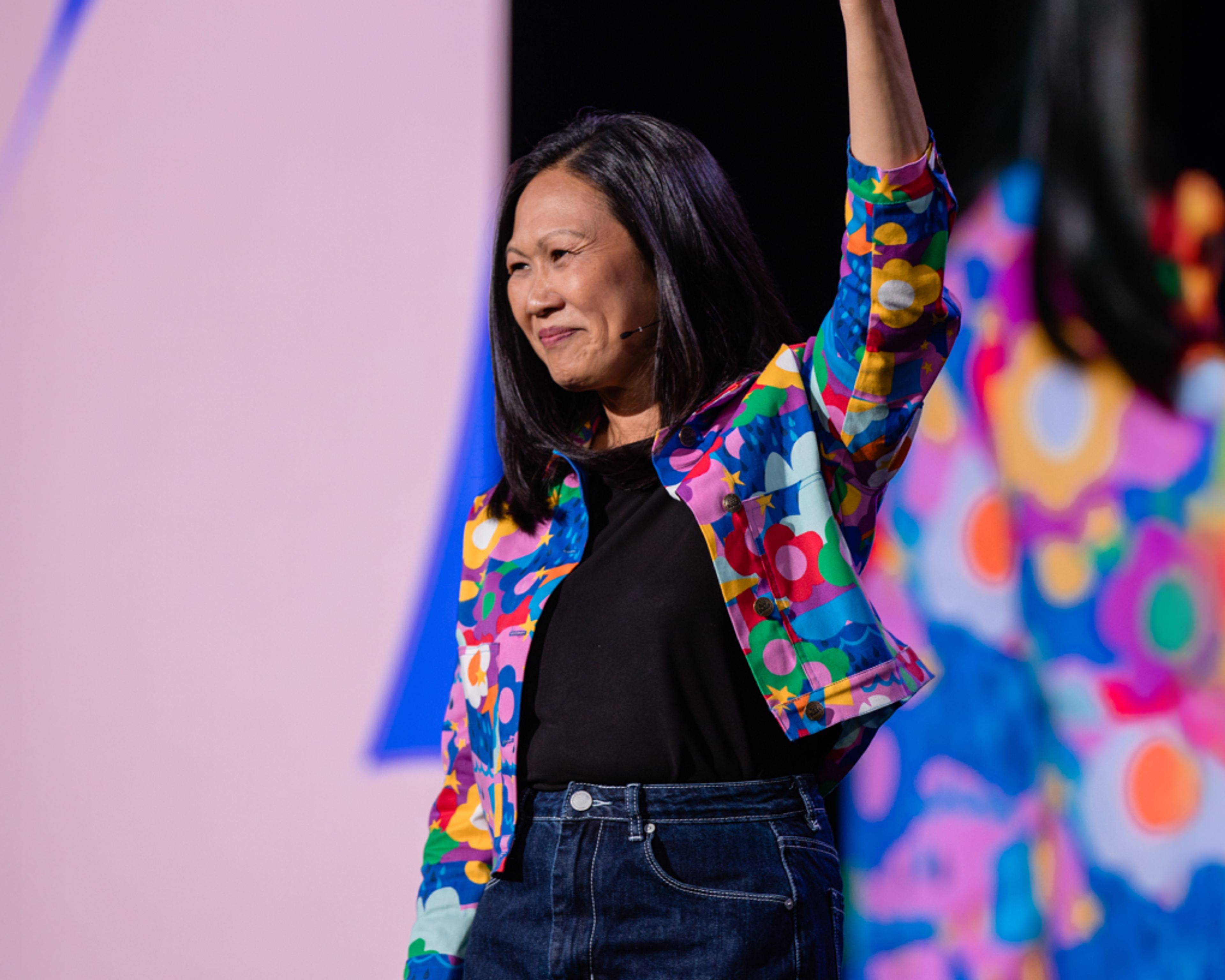 Speaker on stage wearing a bright patterned jacket, smiling with one arm raised.