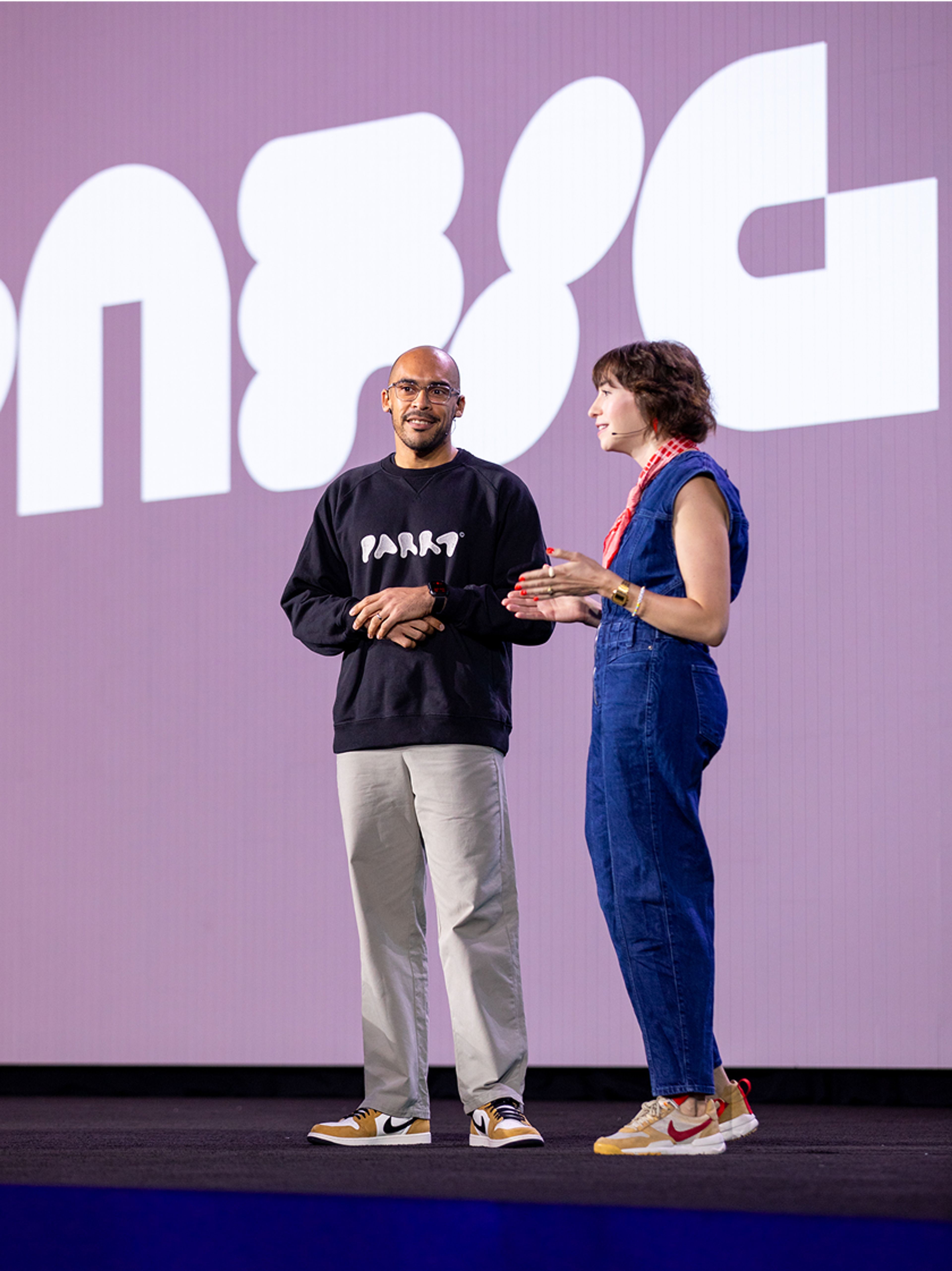 Two speakers on stage at Figma Config 2025, one wearing a black sweatshirt and the other in a denim jumpsuit with a red scarf, engaged in conversation.