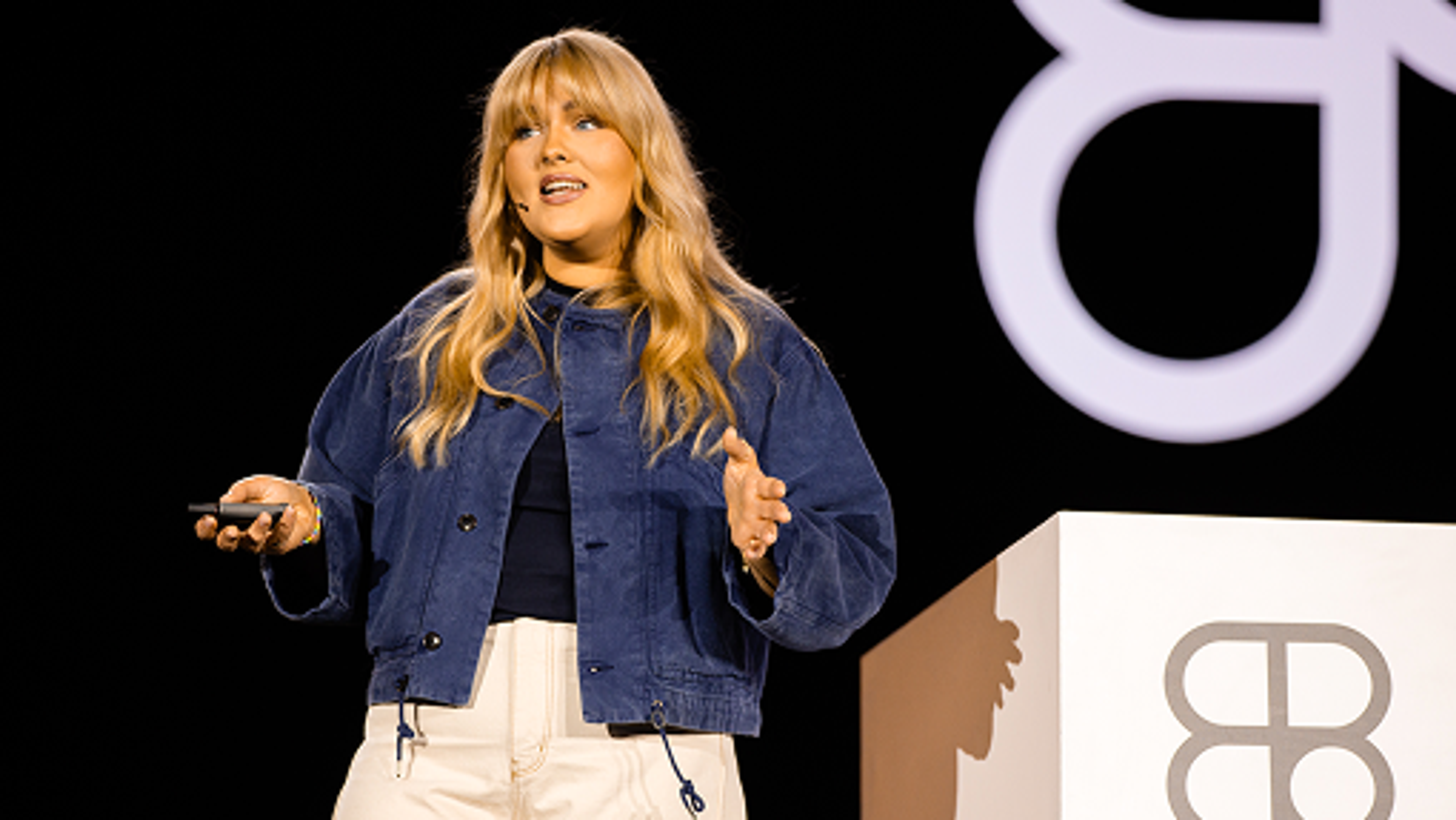 Speaker on stage holding a clicker, wearing a blue jacket and light pants, presenting beside a podium.