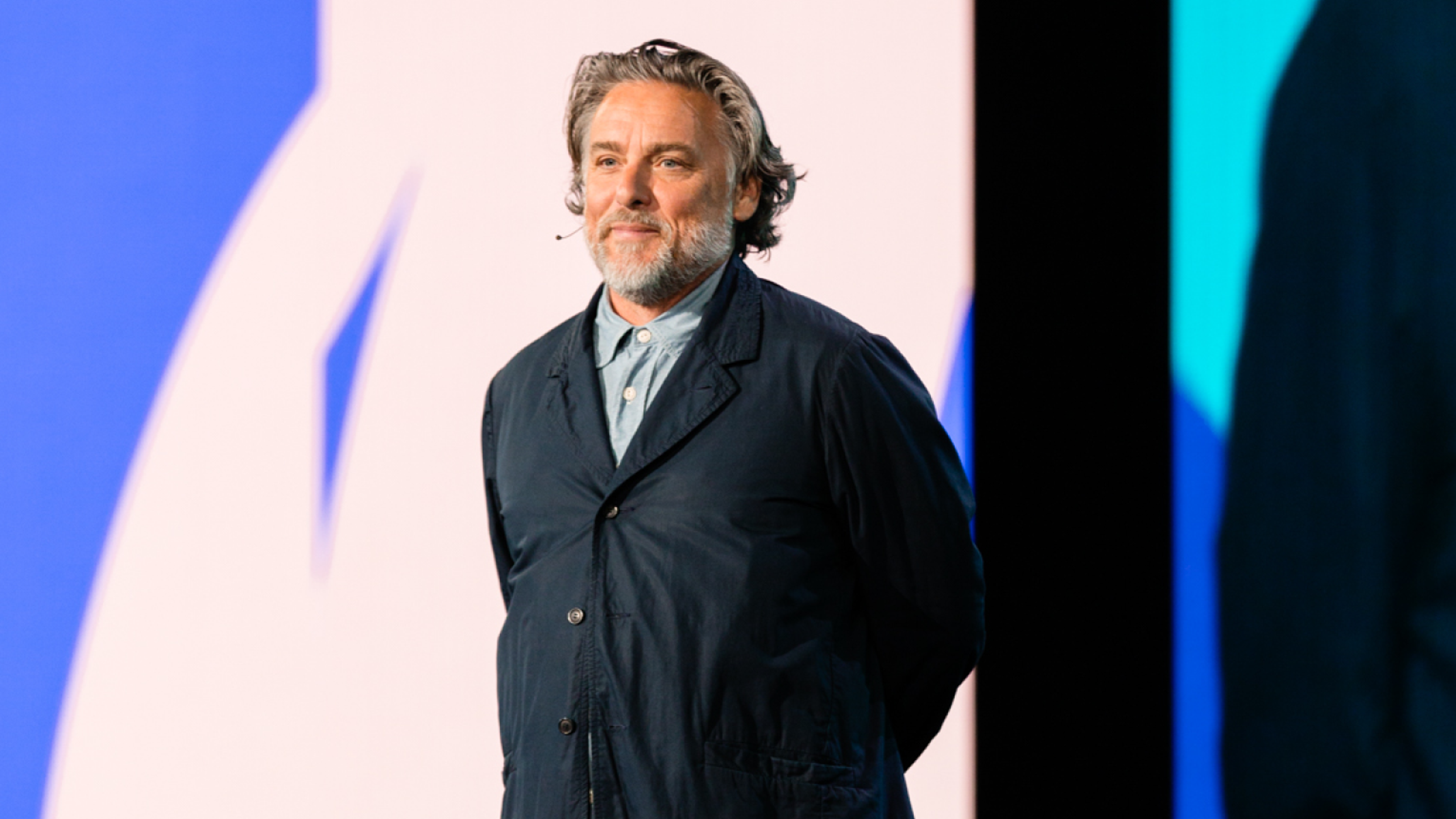 Speaker on stage with their hands behind their back in front of a blue and white background.