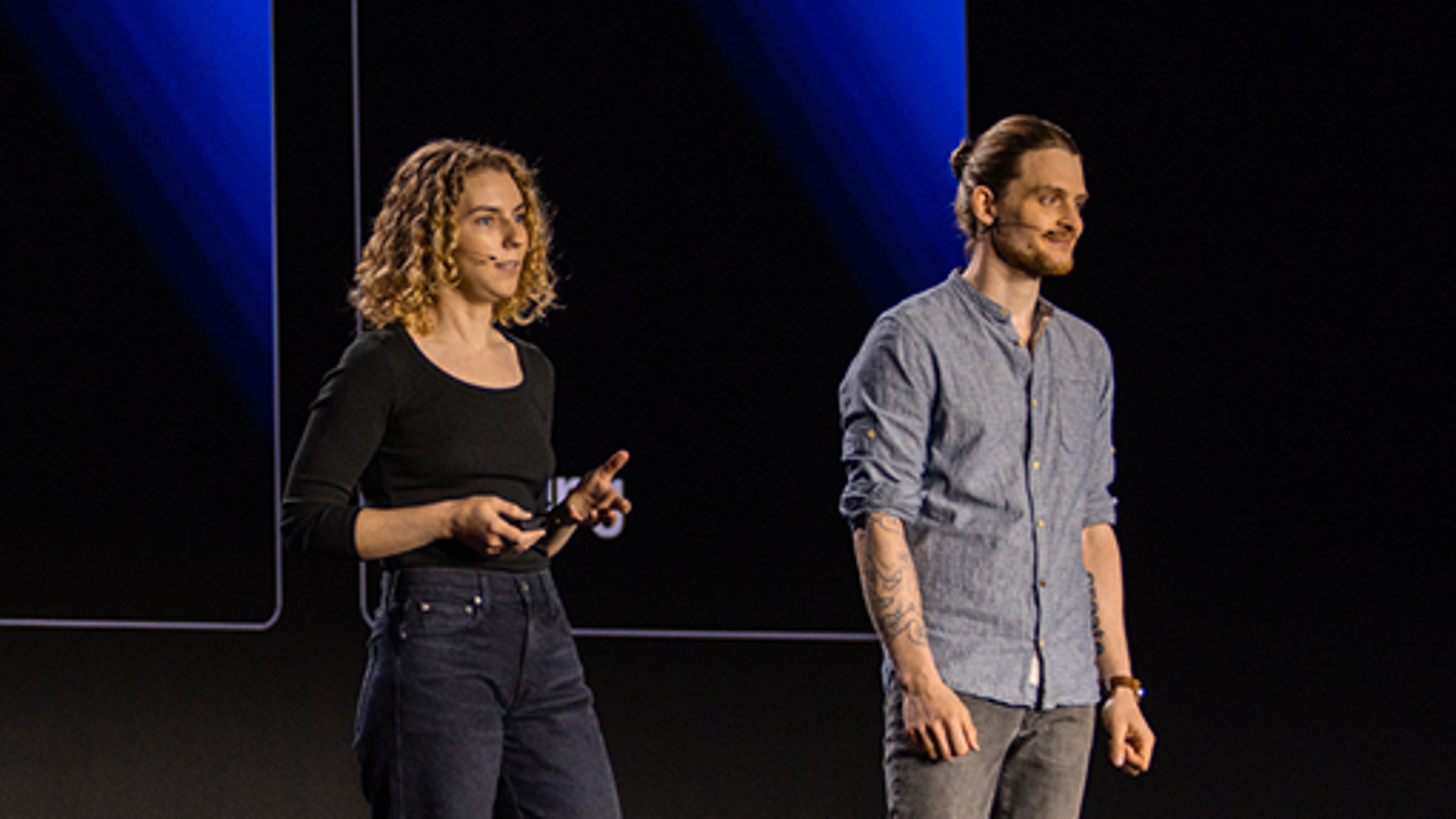 Two speakers on stage, a woman with curly hair wearing a black top and jeans, and a man with tied-back hair, tattoos, and a button-up shirt, both with headsets, presenting in front of a dark backdrop with blue lighting.
