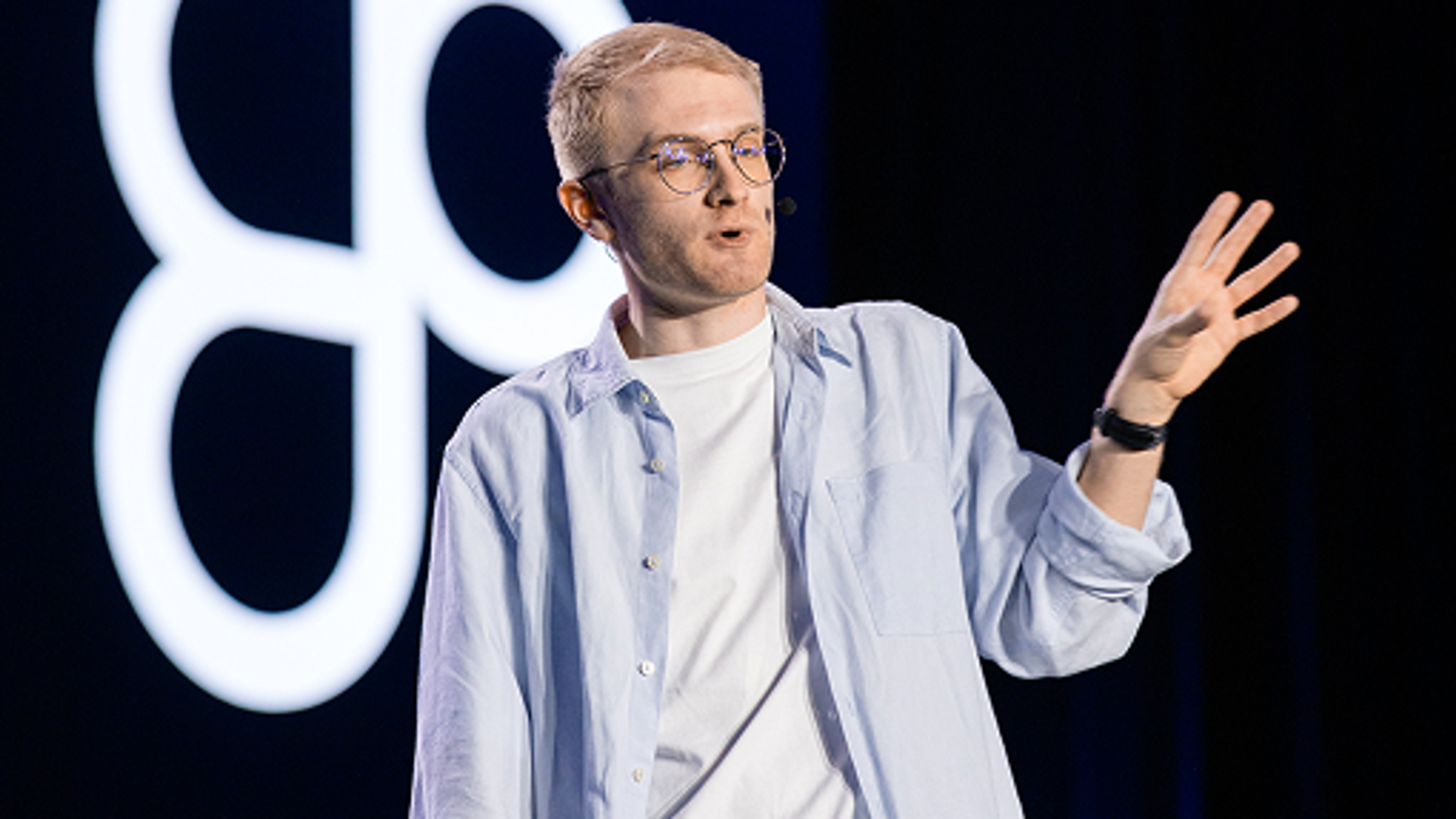 A speaker with short blond hair, glasses, and a headset, wearing a white t-shirt and light blue button-up shirt, speaking on stage against a dark background.