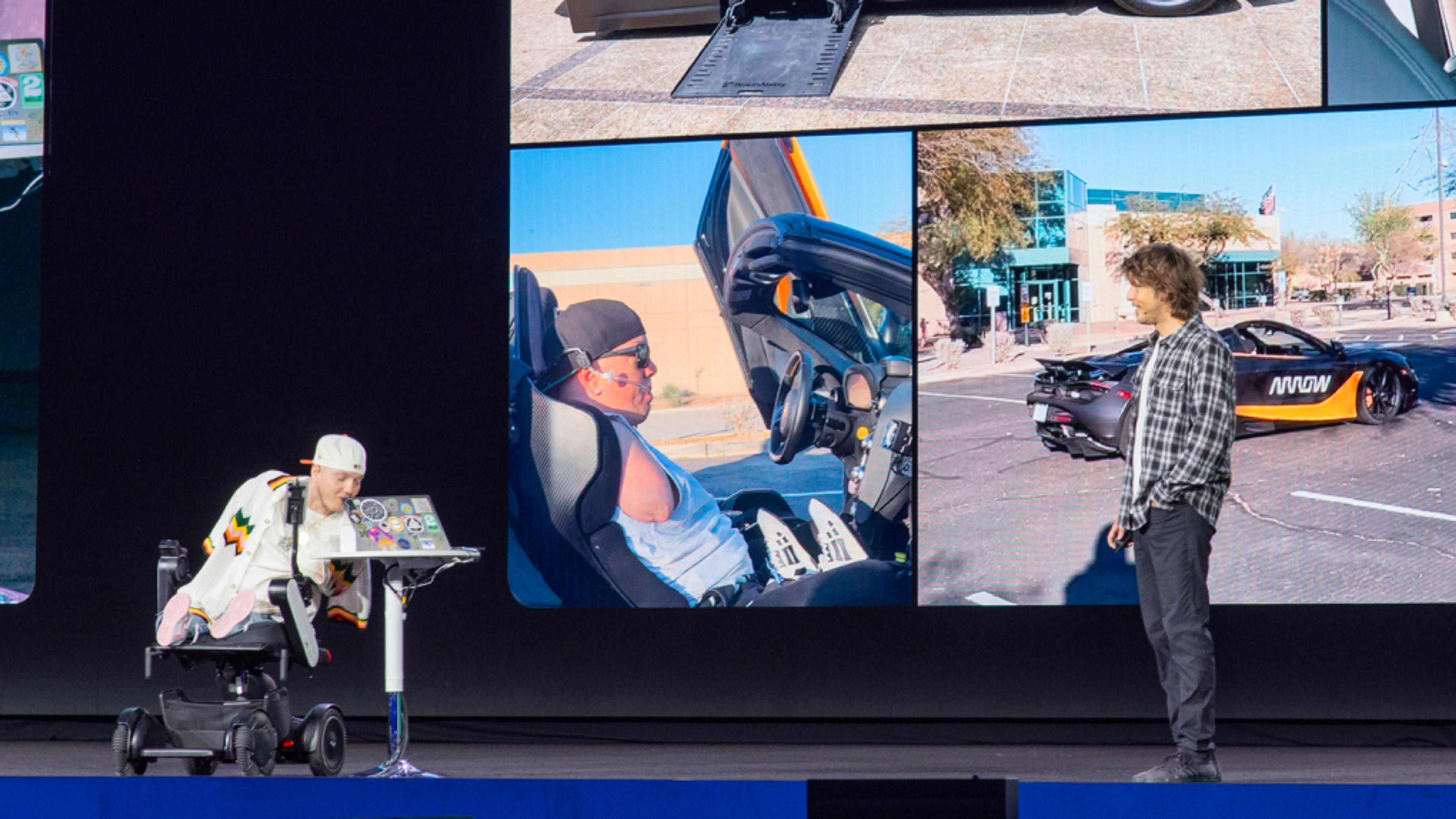 Two speakers on stage with visuals of them driving in a car behind them. One speaker is a person using a wheelchair. The other is standing off to the side.