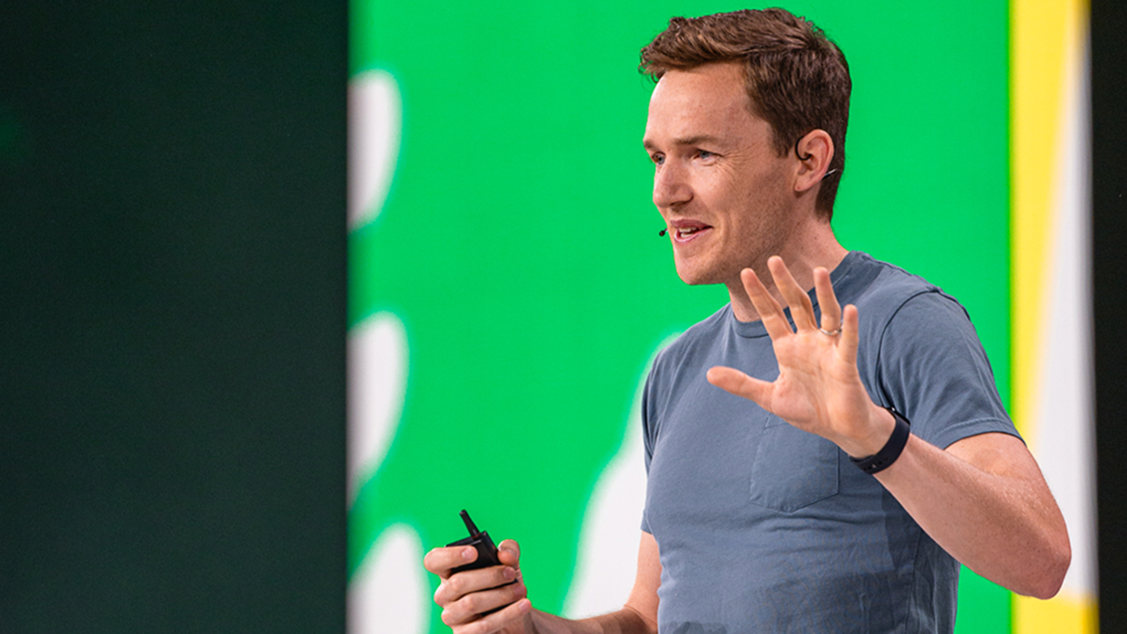 Man speaking on stage with hand up counting to five, green background behind him