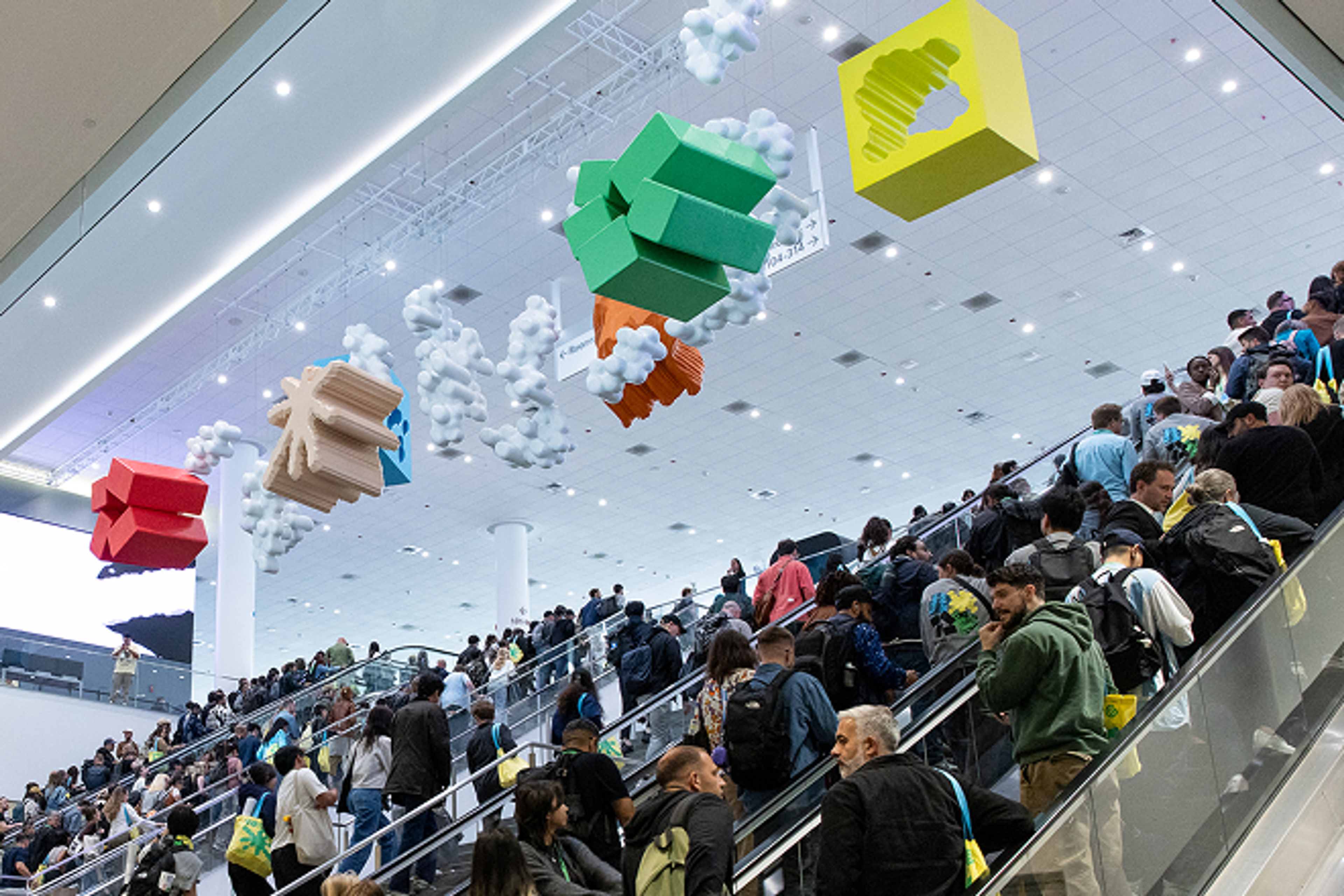 Abstract art hanging from ceiling, art is multi-colored, people are on escalators below