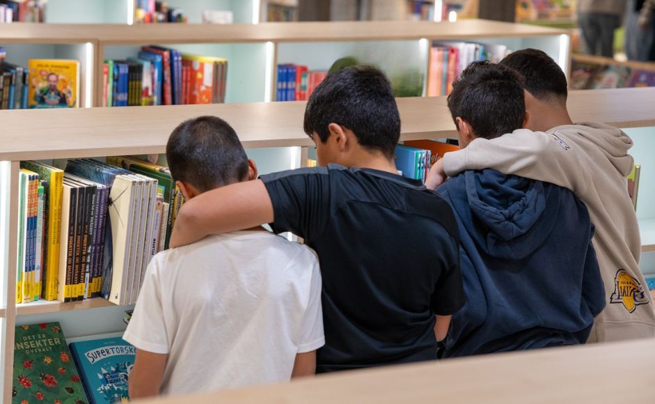 Fire unge gutter står ved en bokhylle på et bibliotek og ser på bøkene. Foto: James Chanika.