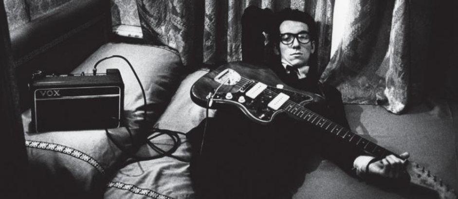 Elvis Costello liggende i seng med gitar på magen