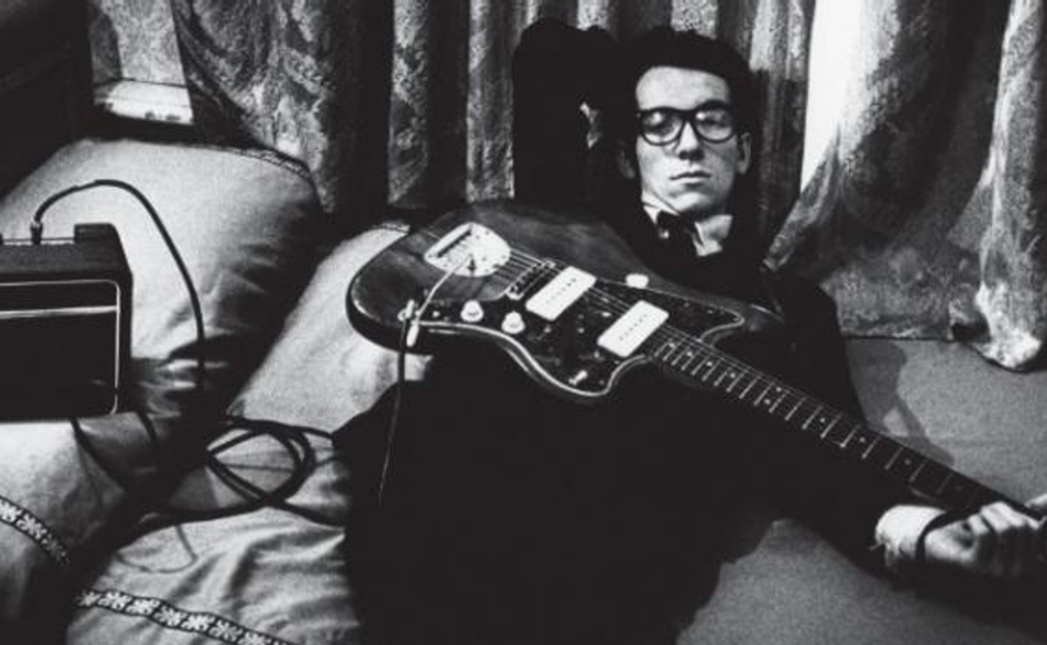 Elvis Costello liggende i seng med gitar på magen