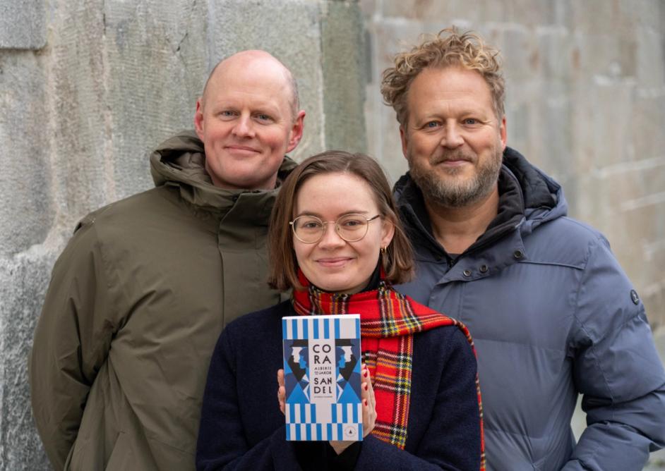 Åsmund Ådnøy, Ruth Stokke Haaland og Tomas Gustafsson med boka Alberte og Jakob av Cora Sandel stående foran en murvegg. Foto: Asbjørn Goa.
