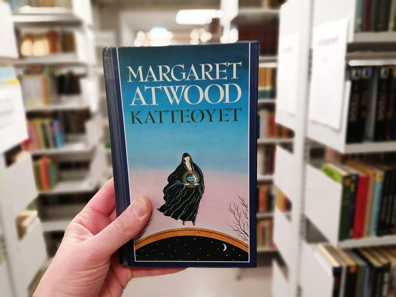 Margaret Atwoods roman Katteøyet holdt foran bokhylle