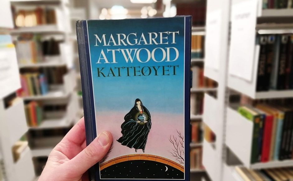 Margaret Atwoods roman Katteøyet holdt foran bokhylle