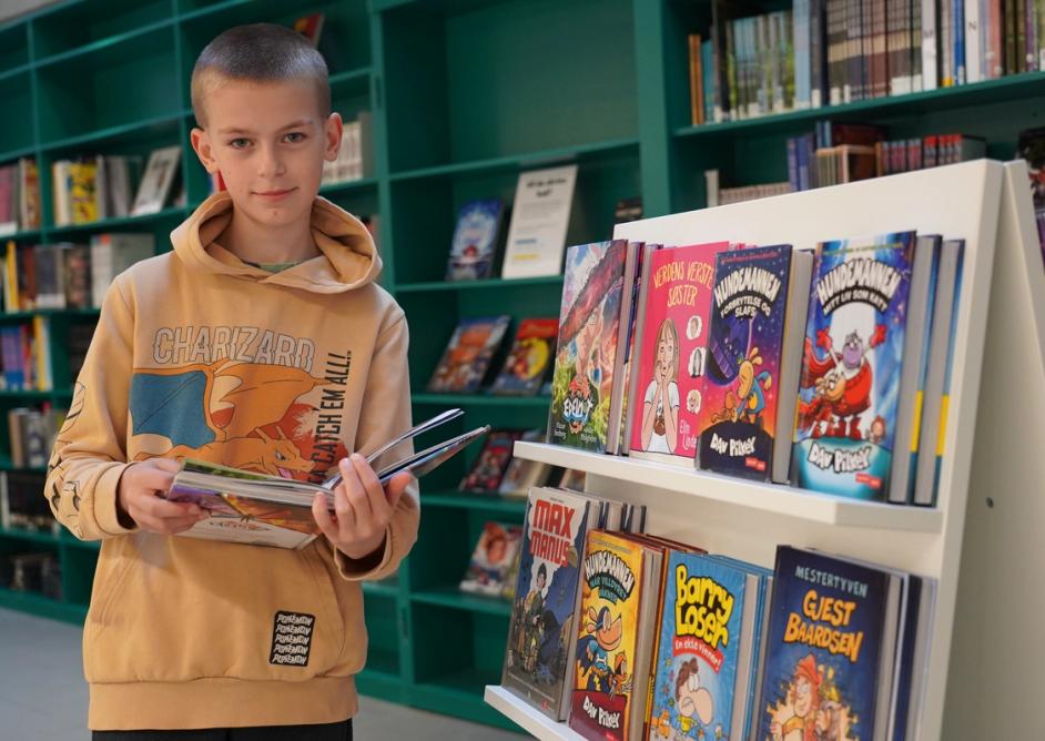 En gutt holder en bok og står ved siden av en bokhylle i biblioteket.
