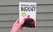 Boka Blodet av Torgrim Eggen holdt foran en vegg