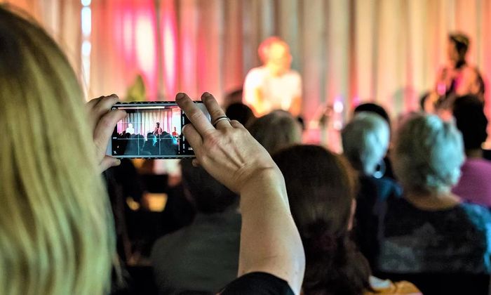 Bilde av arrangementlokale med samtale på scenen og en person tar bilde med mobilen.