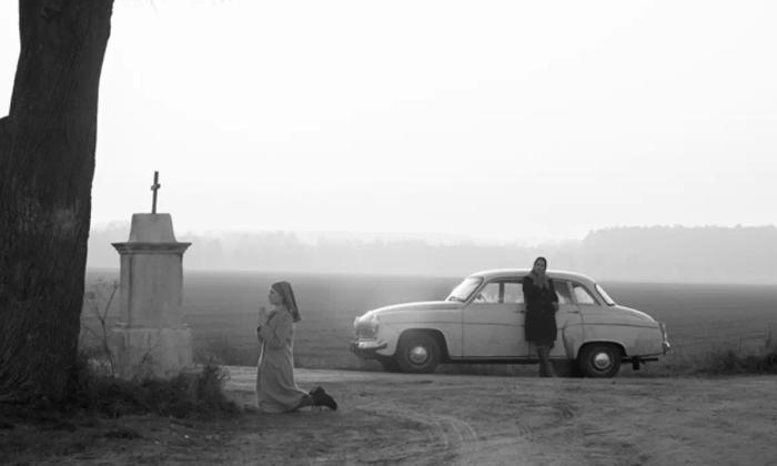 Stillbilde fra den polske filmen Ida fra 2015. Kvinne ved siden av bil kneler foran kors. Svart hvitt.